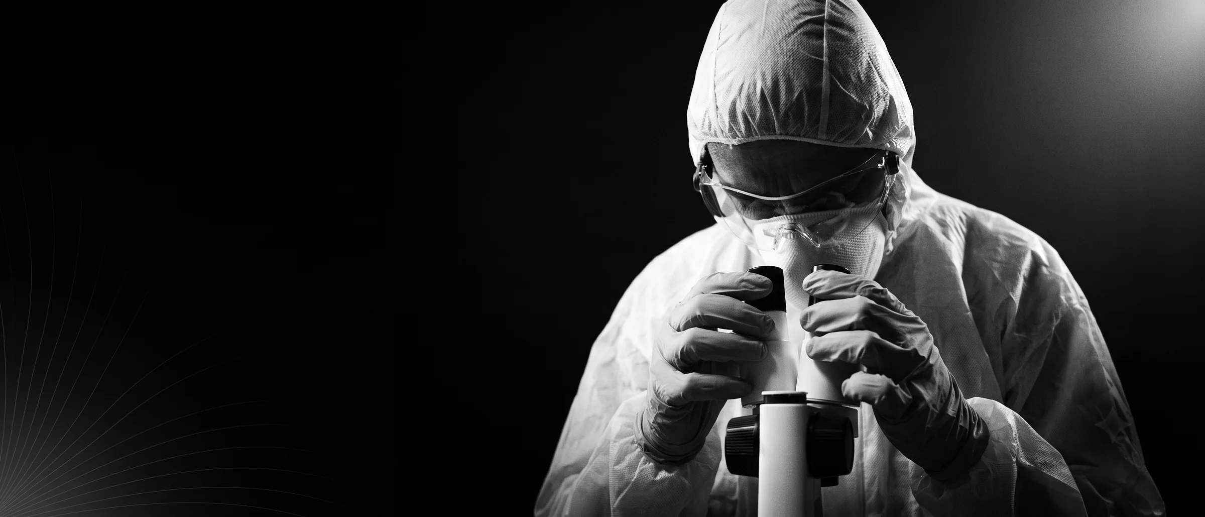 Especialista en traje de laboratorio observando por un microscopio, iluminado con una sola luz dura sobre fondo negro.